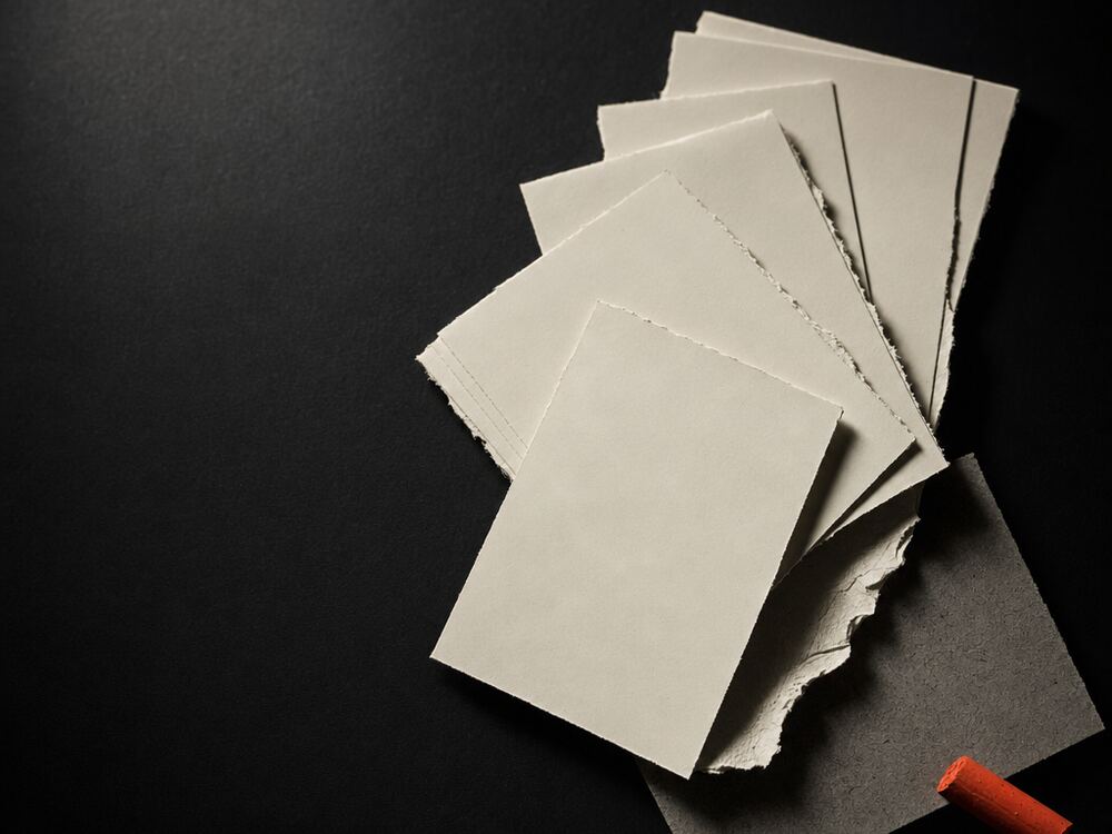 Blank index cards and torn paper strips fanned across a dark studio bench, one marked with a stub of red chalk.