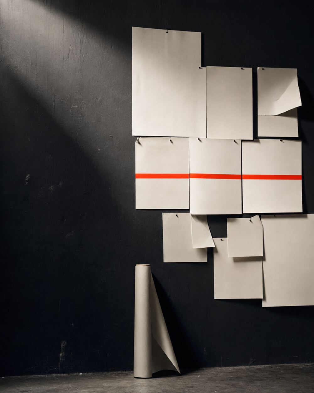 A working studio wall: blank sheets and pale card stock pinned in an uneven grid, corners curling, with a length of red tape running across three of them.
